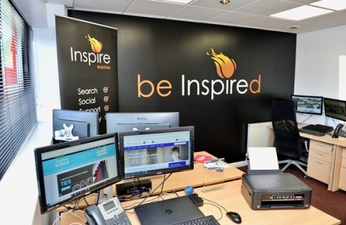 Office workspace at Inspire Digital with multiple computer monitors, telephones, and a wall displaying the 'be Inspired' branding