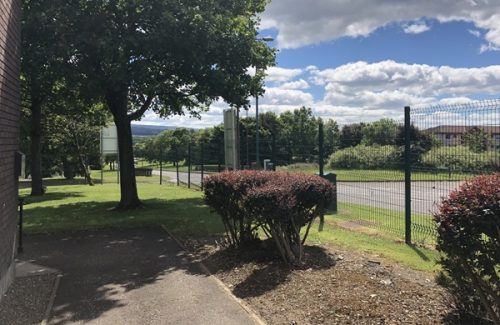 View of the outdoor area near Inspire's Livingston office, showing trees, bushes, a paved path, fencing and partly cloudy blue sky