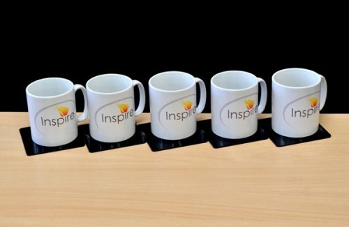 Five white coffee mugs with the Inspire logo lined up on a light wooden office desk