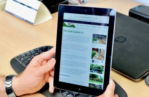 Hands holding an iPad displaying The Good Garden Co website with text and images of gardening services