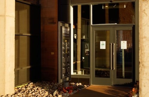 Glass entrance doors of an urban office building with reflective windows and a directory sign beside the door