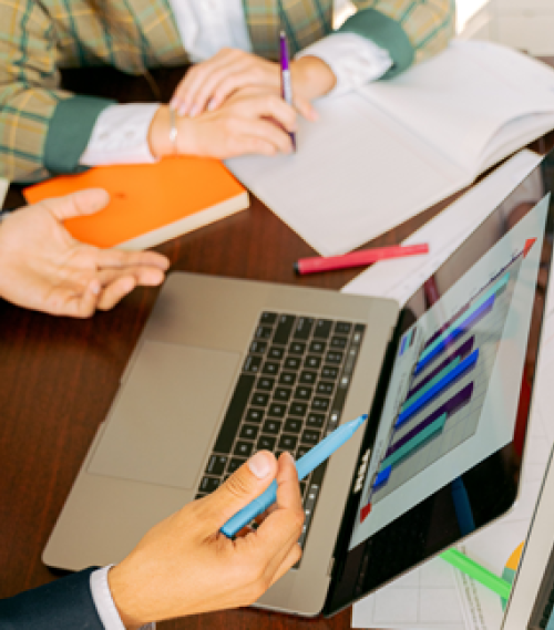 Hands of people in a business meeting with a laptop displaying colourful graphs and open notebooks on the table