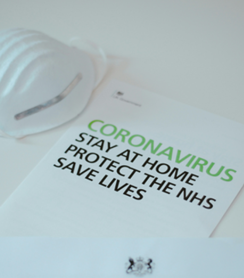 Leaflet titled 'Coronavirus Stay at Home Protect the NHS Save Lives' beside a white face mask on a surface