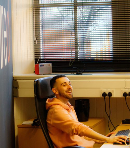 Smiling man in an orange hoodie seated at an office desk while working on a laptop beside a window with black blinds.