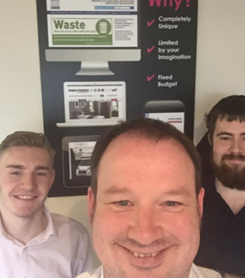 Three men smiling and posing indoors with a promotional poster behind them