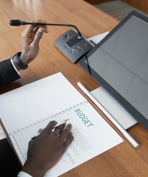 Person at a desk holding a microphone and reviewing a document titled Budget beside a digital screen