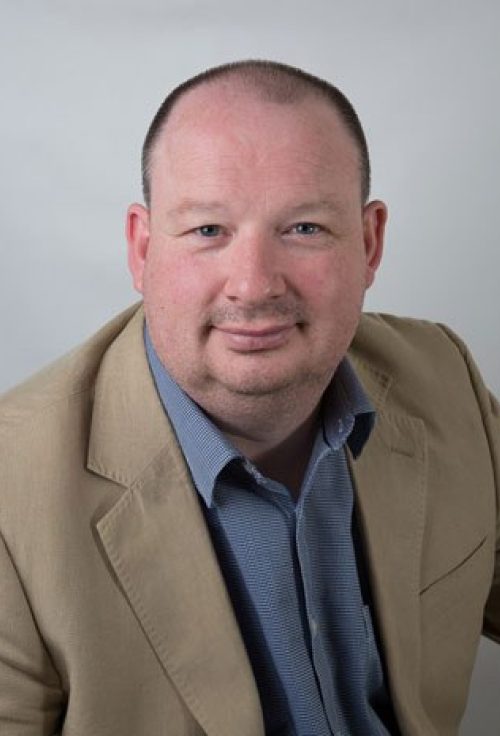Headshot of David Dwyer wearing a beige blazer and blue shirt against a plain background