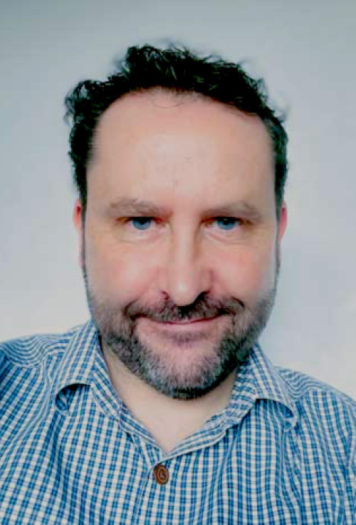 Portrait of a man in a checked shirt with short curly dark hair and a beard, smiling slightly against a plain background