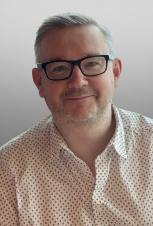 Head-and-shoulders portrait of Martin J Smith wearing black glasses and a patterned white shirt against a light background.