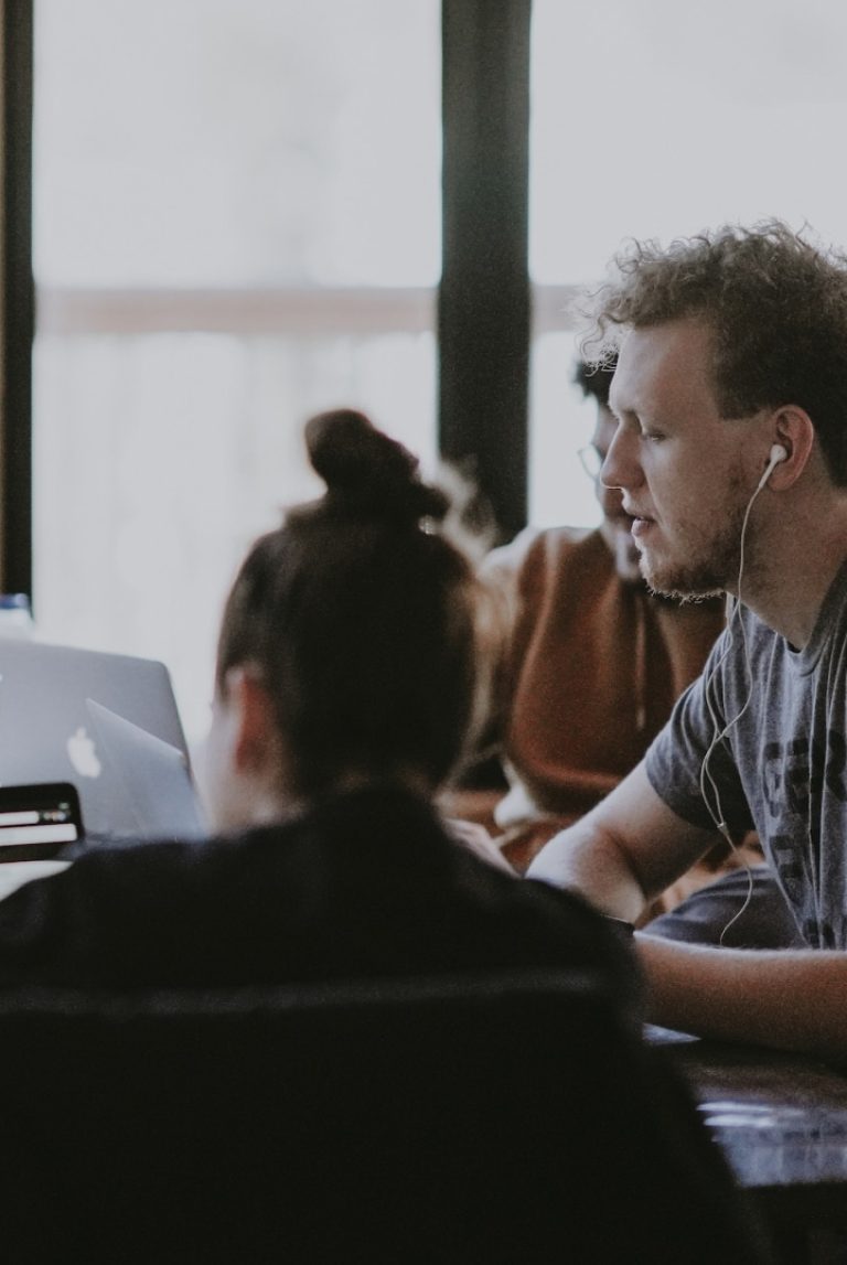 Two people working or collaborating together on laptops in a room with large windows, one person wearing earphones