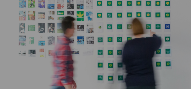 Two people reviewing multiple logo concept designs arranged on a wall during a brainstorming session