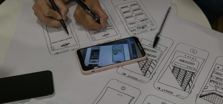 Hands sketching mobile app wireframes on paper with a smartphone displaying digital app designs placed on top