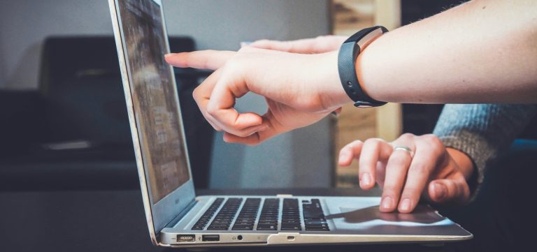 Hand with wristband pointing at laptop screen while another hand types on keyboard
