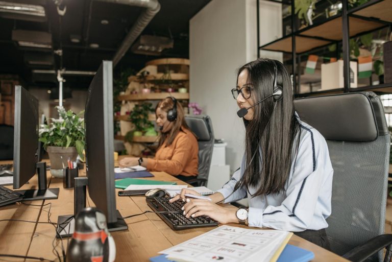 Two women wearing headsets working at computers in a modern office with plants and documents
