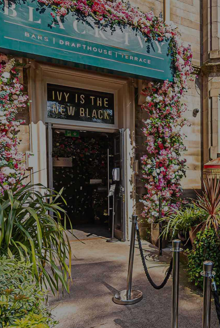 Entrance to Blackivy bar with a green sign above reading 'Bars | Drafthouse | Terrace' and a floral arch around the door